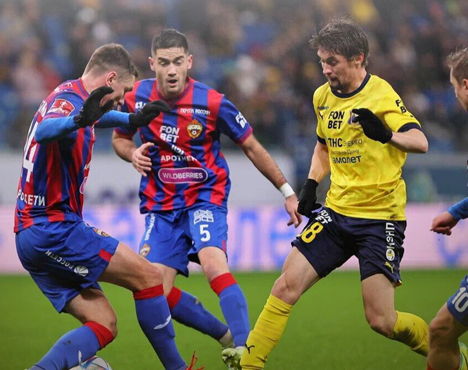 На изображении запечатлен футбольный матч: игроки в синей и красной форме противодействуют игроку в желтой форме. Действие происходит на поле, футболисты сосредоточены на борьбе за мяч