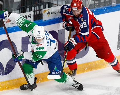 Хоккеисты в зелено-белой и красно-синей форме активно борются за шайбу у борта на льду. Игрок в красно-синем пытается завладеть шайбой, затиснув соперника к борту. Напряженная игра на арене