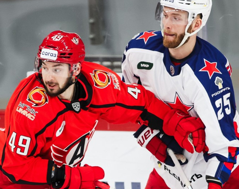 На изображении два хоккеиста в игре. Один в красной форме и шлеме, с номером 49, другой в бело-синей форме с номером 25. Они находятся в борьбе за шайбу на льду, каждый сосредоточен на игре