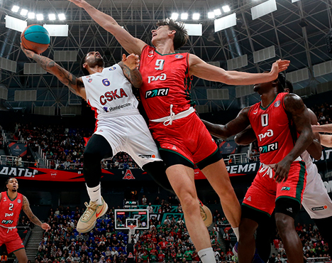 На изображении баскетбольный матч. Игрок в белой форме пытается забросить мяч в кольцо, противостоя игроку в красной форме, который стремится блокировать бросок. На заднем плане трибуны с болельщиками