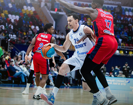 На изображении запечатлён момент баскетбольного матча. Игрок в белой форме ведёт мяч, пытаясь обойти защитника в красной форме. На заднем плане видны сидящие зрители