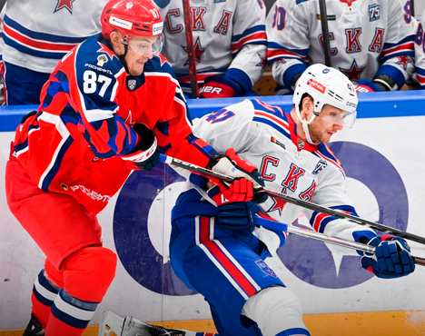 На изображении два хоккеиста борются за шайбу у борта. Один в красной форме, другой в белой с символикой СКА. На заднем плане видна скамейка с игроками в форме СКА