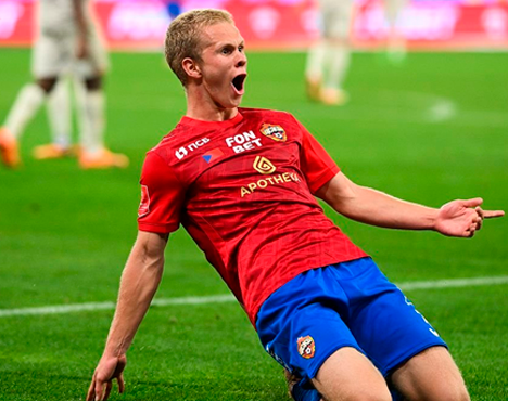 Футболист в красной форме с эмблемой и логотипами спонсоров празднует гол, совершая эффектное слайд движение на коленях по зелёному футбольному полю. На заднем плане видны другие игроки