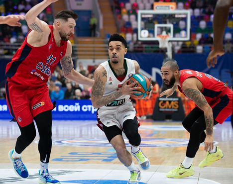 На изображении трое баскетболистов в спортивной борьбе на площадке. Игрок в белой форме пробивается к кольцу между двумя игроками в красной форме. Обстановка напряженная, задний план с размытыми зрителями